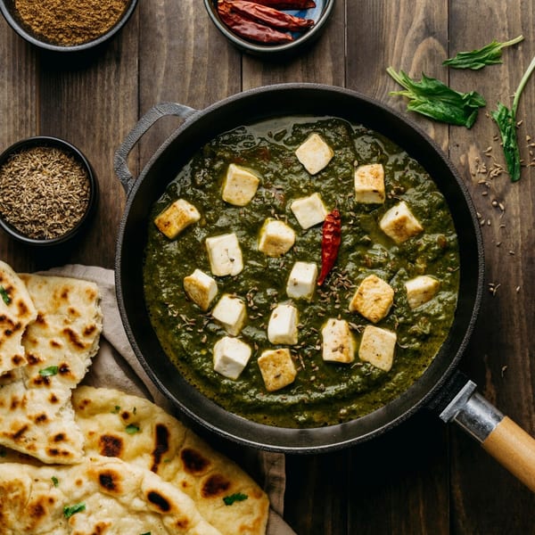 Palak Paneer in a black pan with naan bread, spices, and spinach on a wooden table.