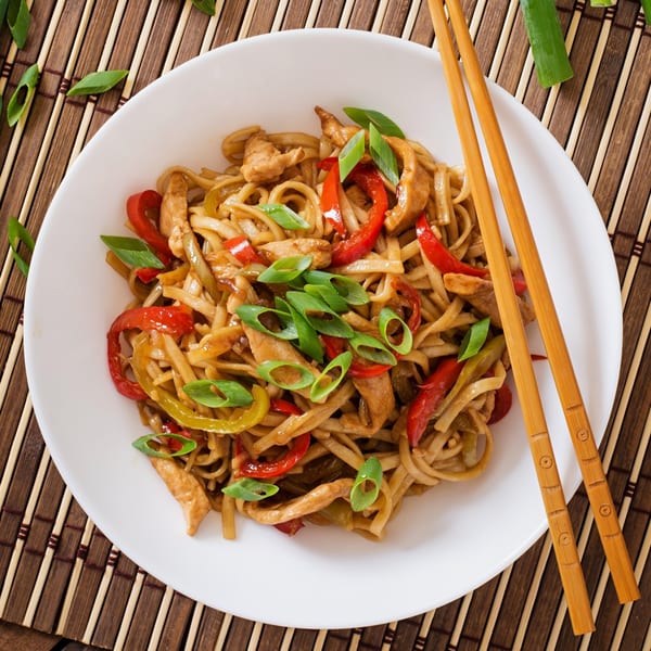 Chicken chow mein with red and green bell peppers, and green onions on a white plate with chopsticks.
